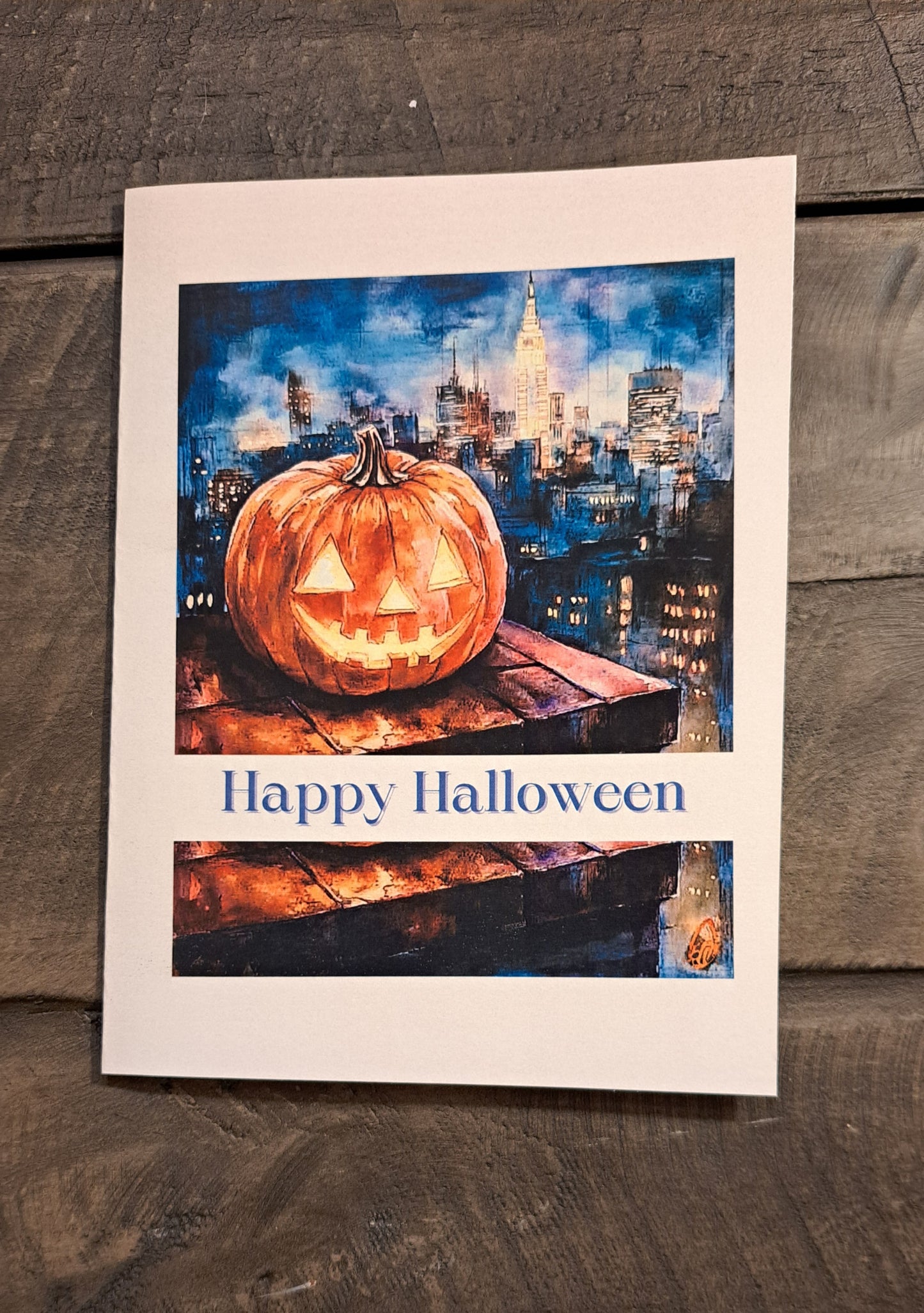 Halloween card with a carved, smiling pumpkin on a rooftop overlooking a city at night, with the message “Happy Halloween”
Image Description: A greeting card showing a carved pumpkin on a rooftop with a cityscape at night. The cover says “Happy Halloween,” and the inside reads “Sending you pumpkin spice wishes and delightful treats.”